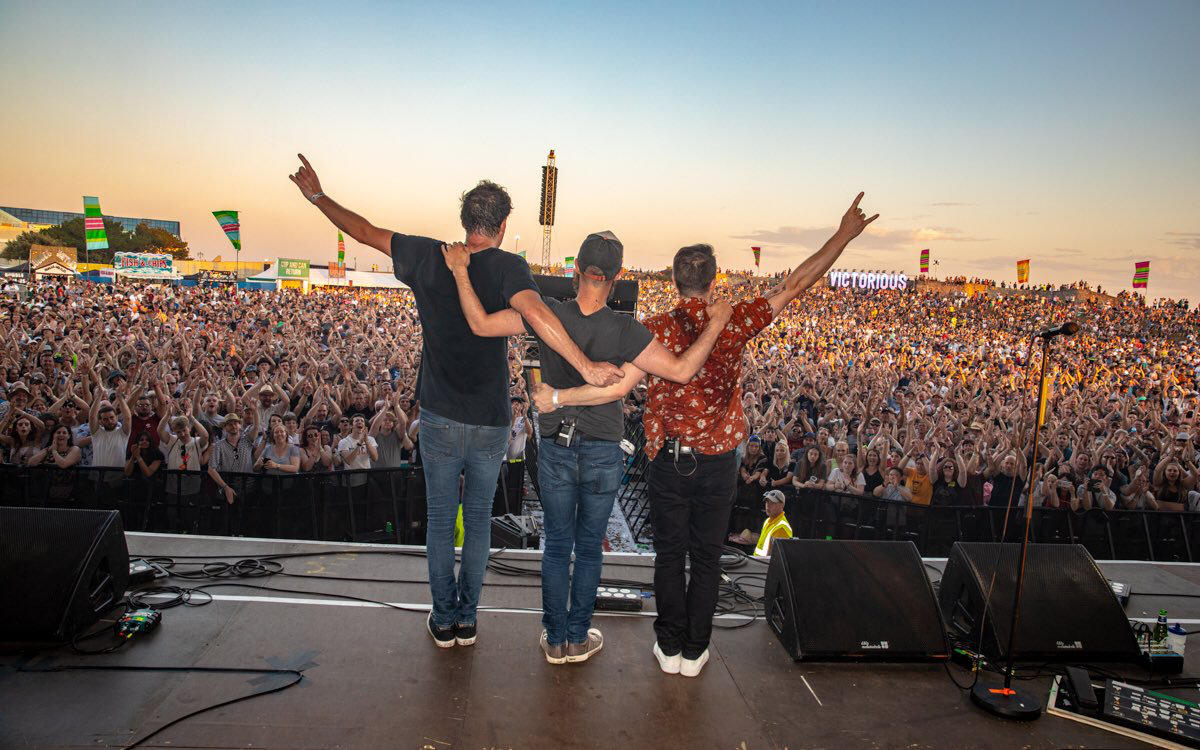 The three members of Ash arm in arm face a festival crowd with their backs to the camera.
